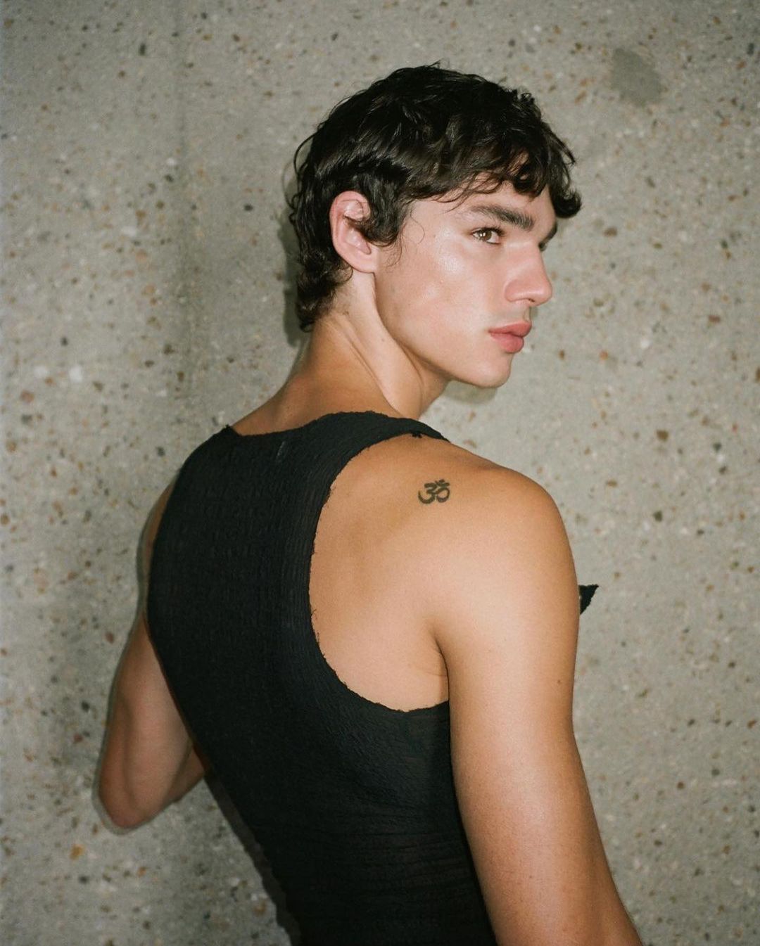 Portrait of a young adult male with dark hair, wearing a black tank top, looking over his shoulder in front of a textured gray wall.