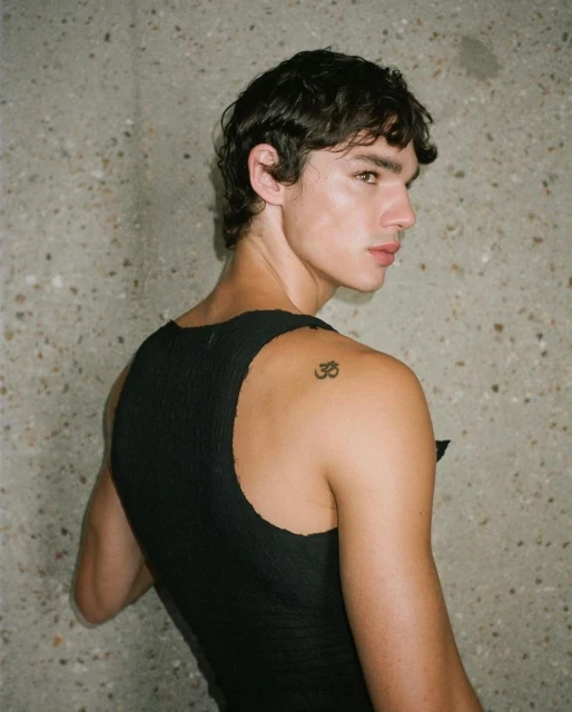 Portrait of a young adult male with dark hair, wearing a black tank top, looking over his shoulder in front of a textured gray wall.