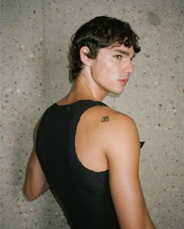 Portrait of a young adult male with dark hair, wearing a black tank top, looking over his shoulder in front of a textured gray wall.