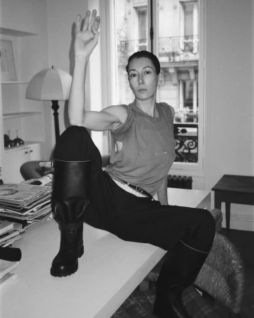Monochrome portrait of a woman seated on a desk wearing a sleeveless top, dark trousers, and tall boots, photographed in Paris.