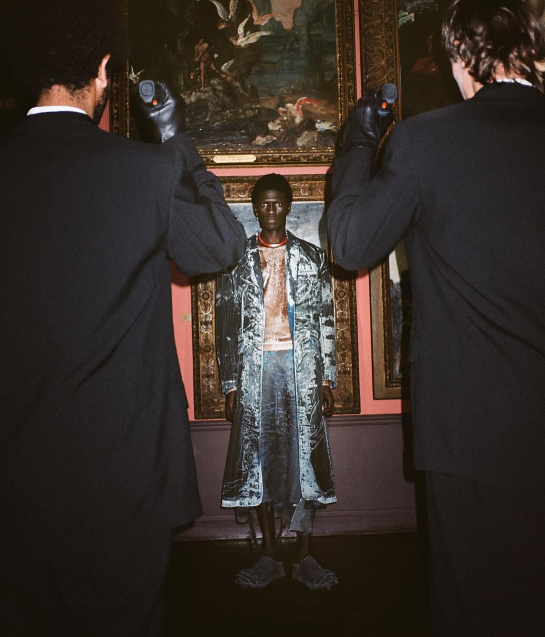 A black model stands between two men holding light meters, framed by ornate paintings in an art gallery, under soft, directional lighting.
