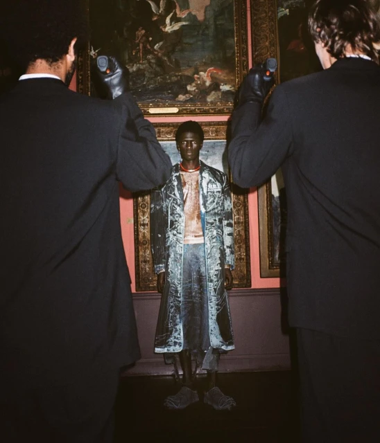 A black model stands between two men holding light meters, framed by ornate paintings in an art gallery, under soft, directional lighting.