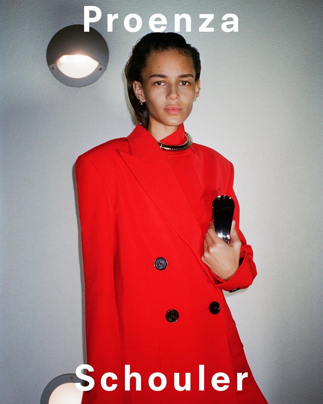 Elegant adult female model in a vibrant red blazer and gold choker poses in a minimalist indoor setting with sleek hair and fair skin.