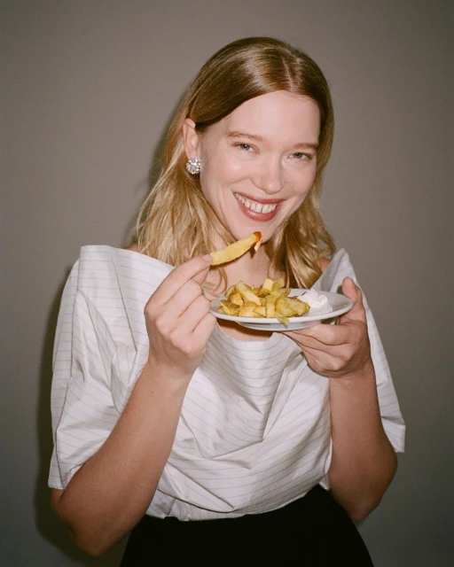 Smiling woman in a white shirt holds a plate of French fries and a condiment. She is eating a single fry with ketchup.