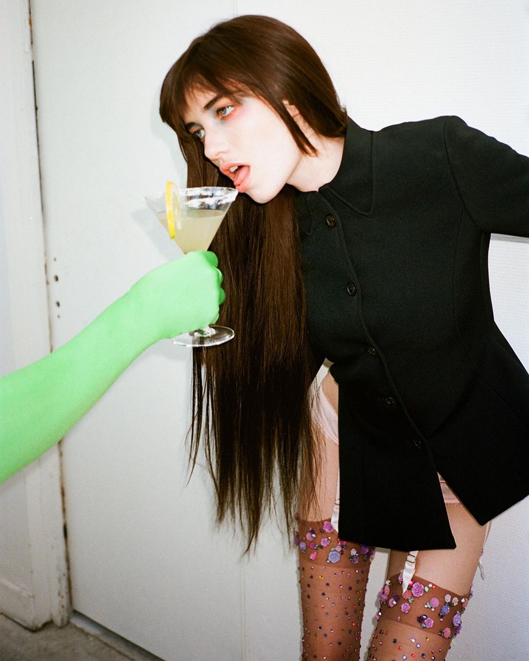 A young adult woman with long brown hair drinks a martini from a green gloved hand in a studio setting, wearing a dark jacket and hosiery.