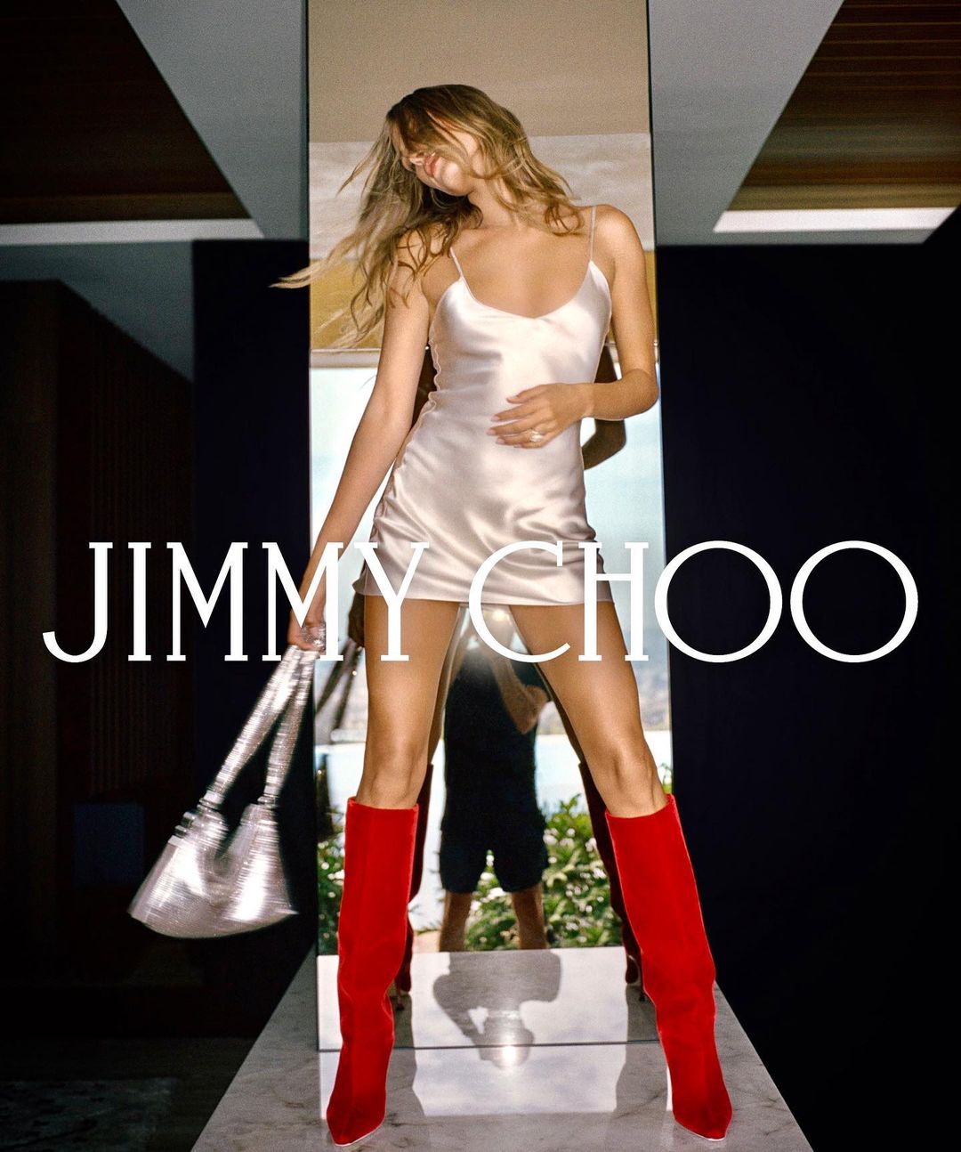A woman poses in a champagne-colored dress, silver purse, and red boots, highlighted by the Jimmy Choo logo and indoor setting.