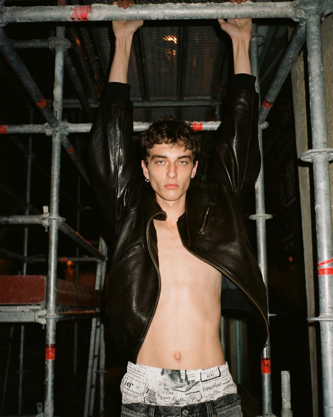 A young adult male model hangs on metal scaffolding wearing a black leather jacket and patterned jeans, shot in medium framing indoors.