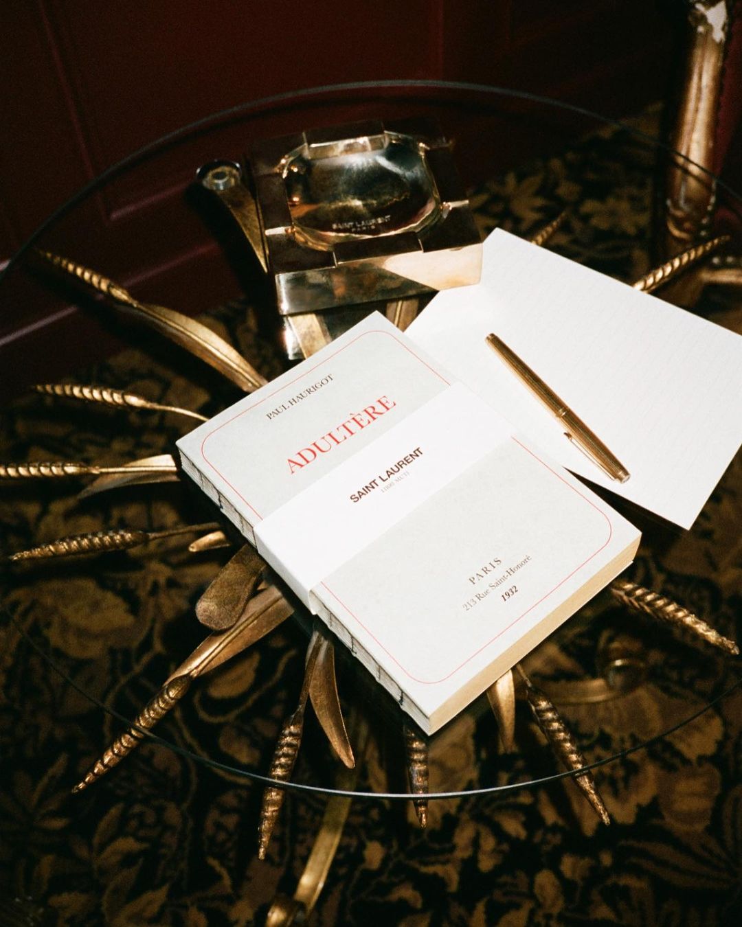 Image features a book titled 'Adultere' by Paul Bourget and 'Saint Laurent' on a glass table with a gold pen and metal ashtray.