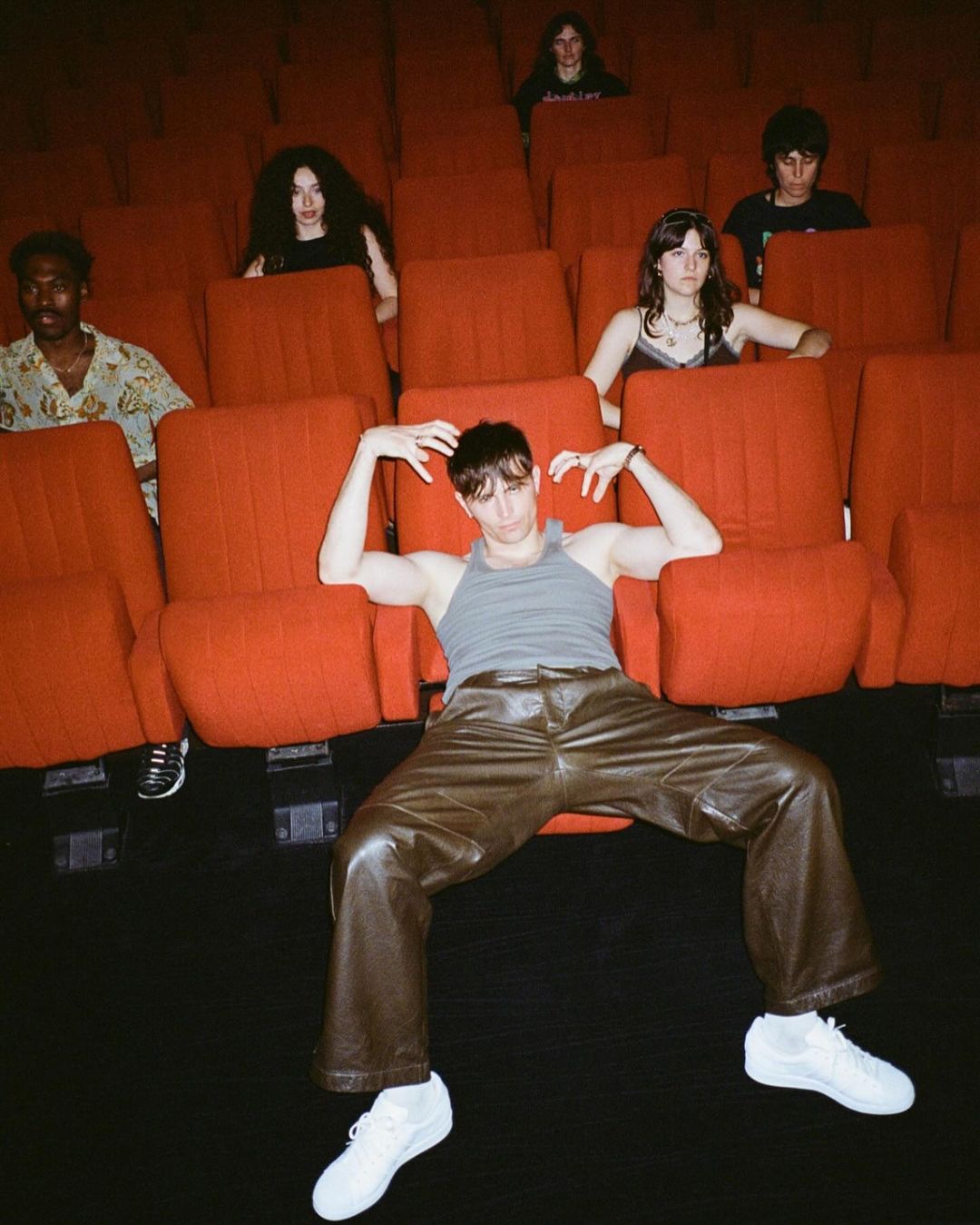 Person in a gray tank top and brown leather pants posing in a movie theater with a group of people sitting in the red seats behind.