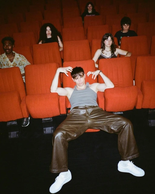 Person in a gray tank top and brown leather pants posing in a movie theater with a group of people sitting in the red seats behind.