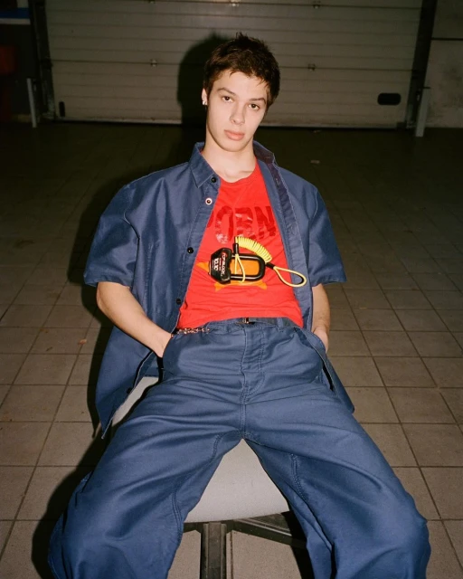 A young man sits on a chair wearing a blue shirt and pants with a red graphic tee in an indoor setting.