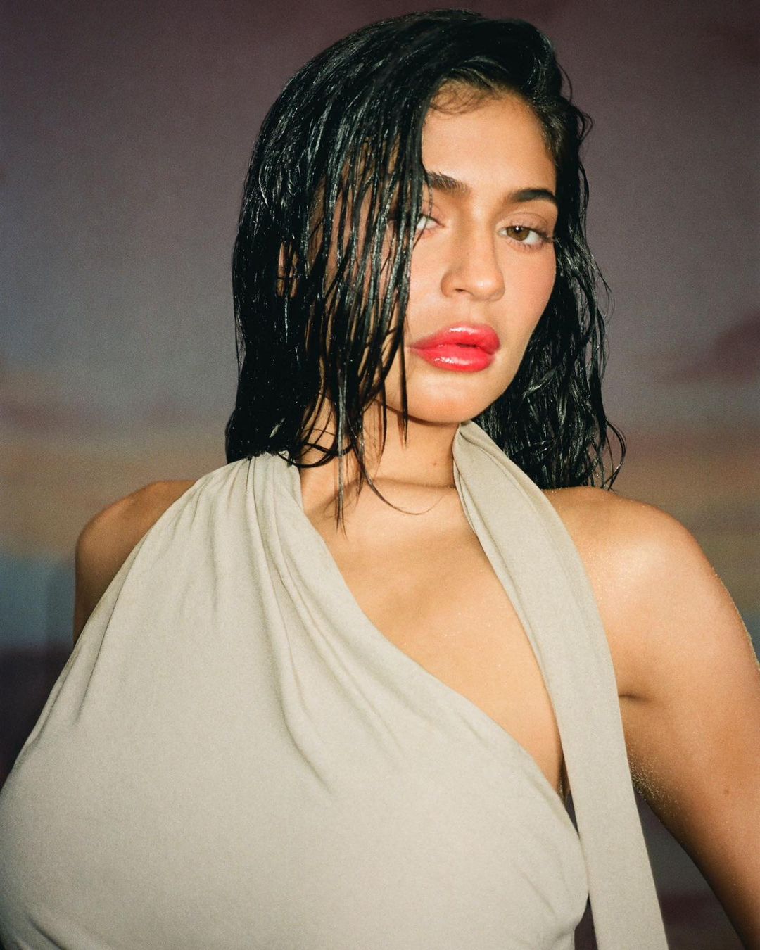 Close-up portrait of a woman with wet dark hair, wearing a neutral-toned dress, and vibrant red lipstick against a neutral backdrop.