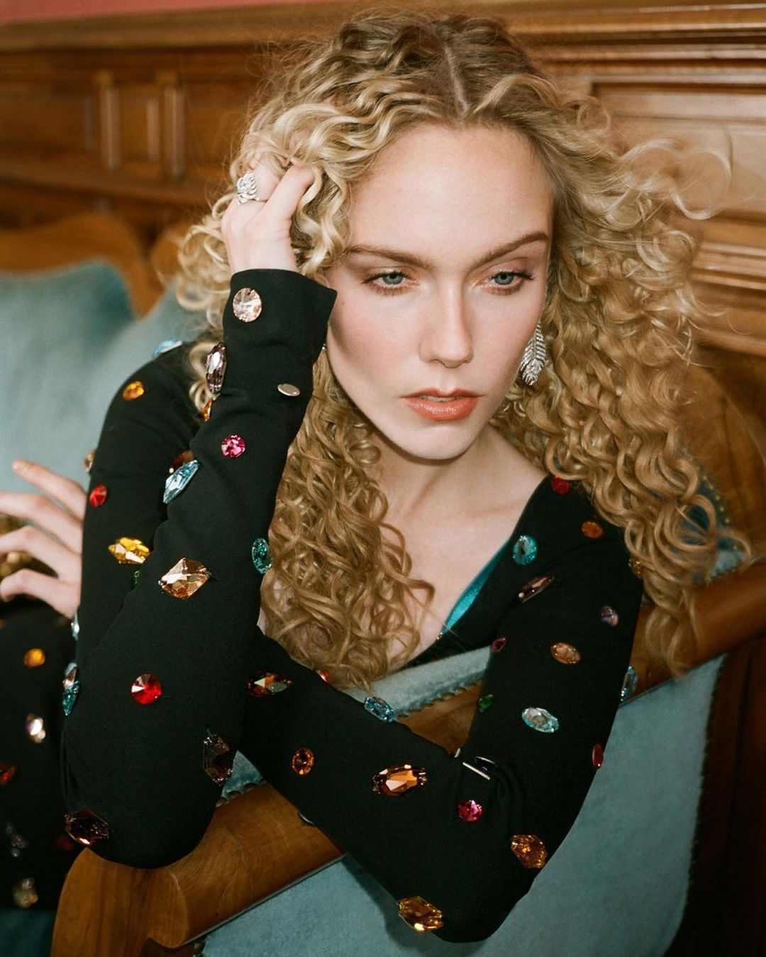 Portrait of a fair-skinned woman with curly blonde hair wearing a bejeweled black dress, sitting on a blue sofa in a warm interior.