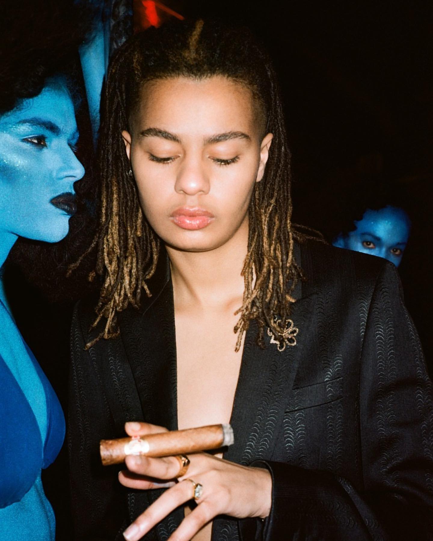 A young person in a blazer holding a cigar, posed with two blue-skinned people in the background against a dark, smoky backdrop.