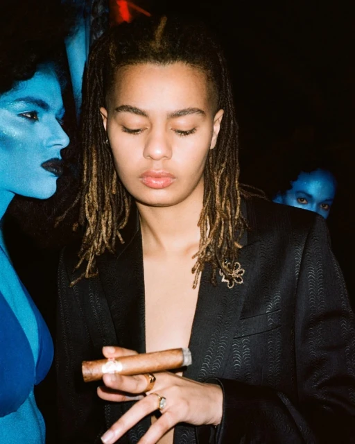 A young person in a blazer holding a cigar, posed with two blue-skinned people in the background against a dark, smoky backdrop.