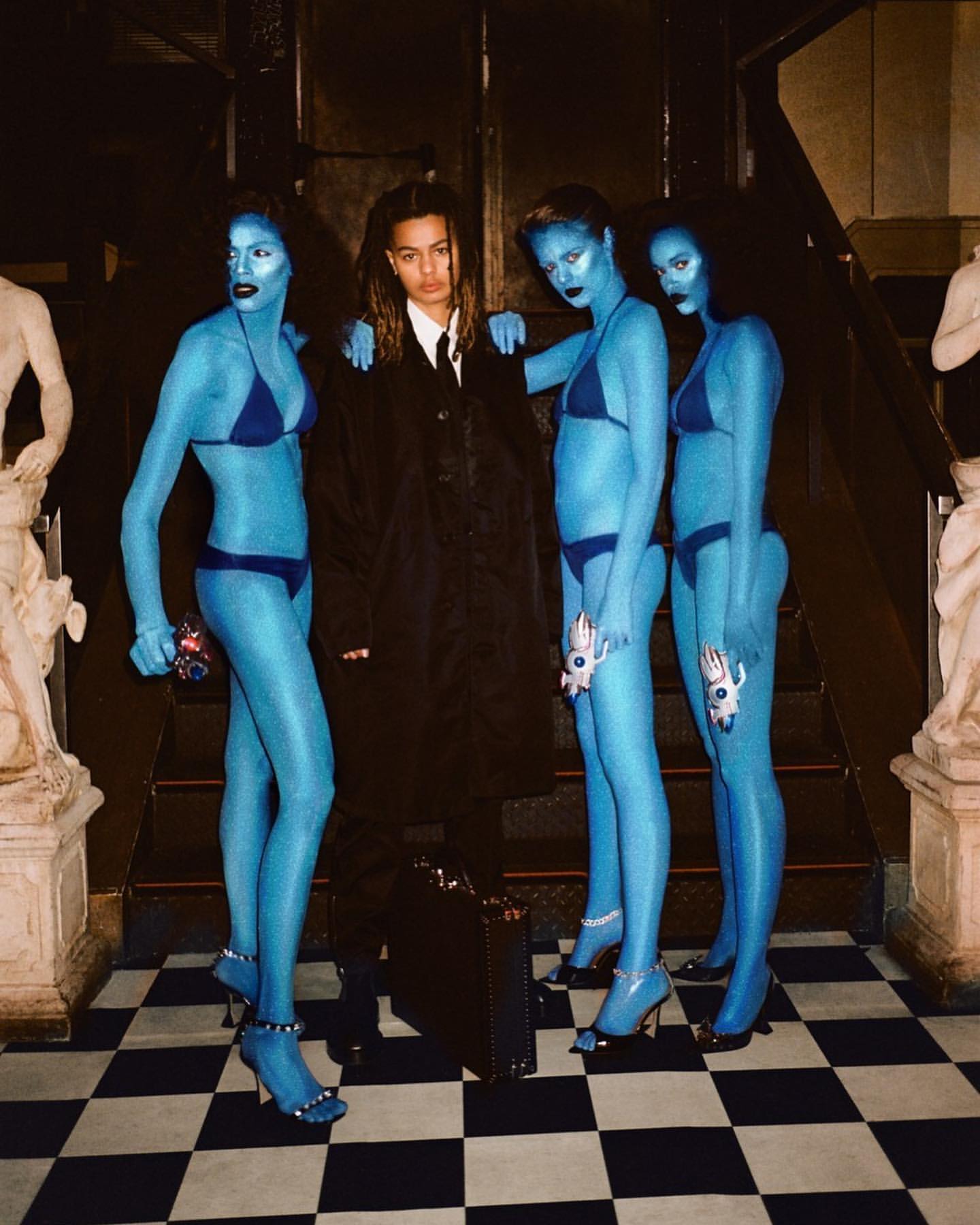 Person in suit surrounded by three figures with blue skin and swimsuits, posing on a black and white checkerboard floor indoors.