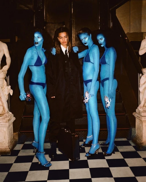 Person in suit surrounded by three figures with blue skin and swimsuits, posing on a black and white checkerboard floor indoors.