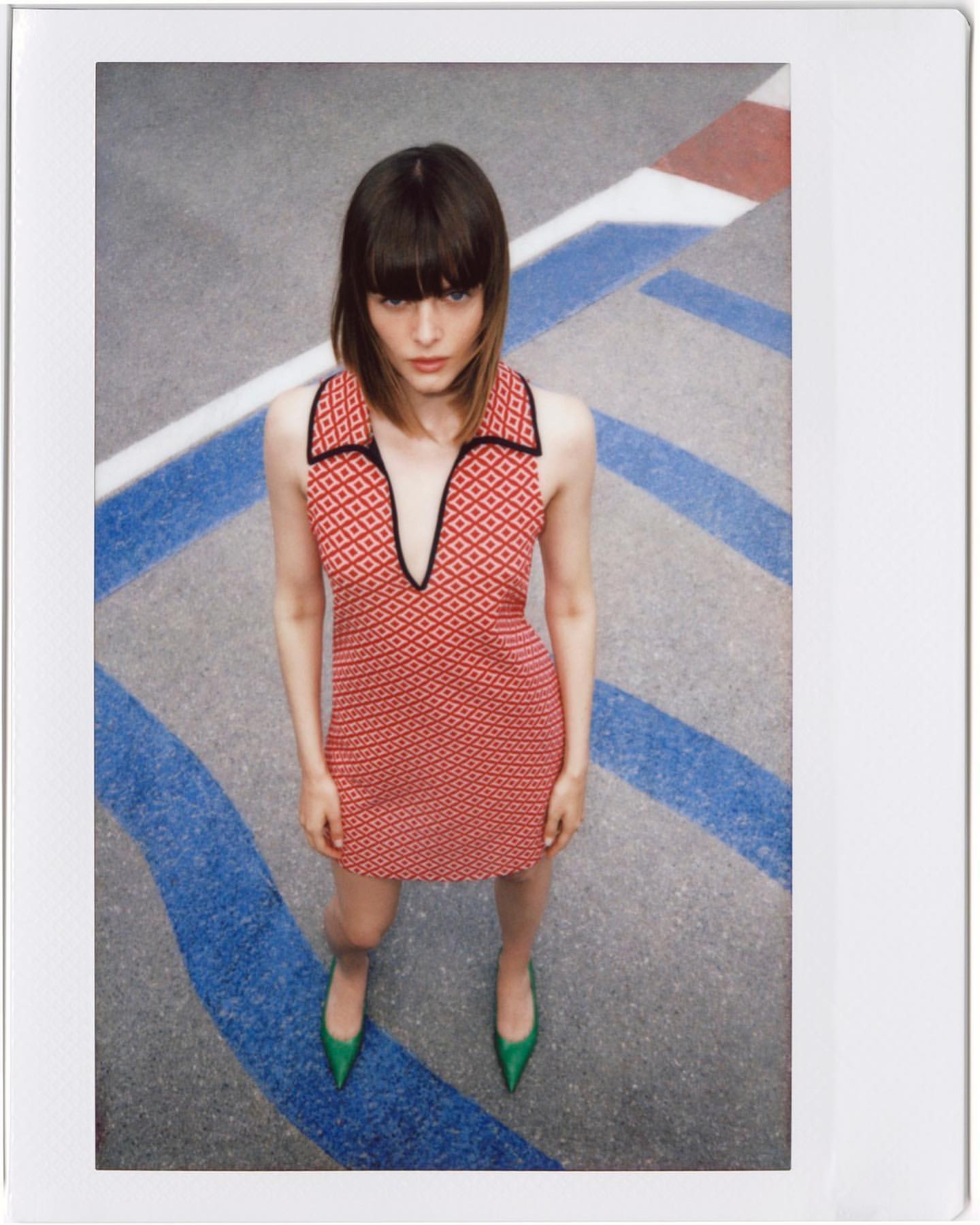 Woman in patterned mini dress and green heels stands on parking lot surface with blue and white painted lines, shot from high angle.