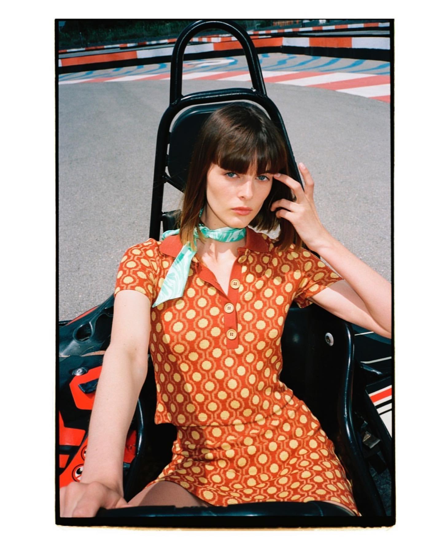 A young woman in a patterned dress sits in a black go-kart on a race track, looking at the camera with a neutral expression.