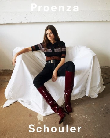 Woman seated on a white-sheeted sofa wearing a striped knit top, dark pants, and glossy burgundy boots, photographed by Pierre-ange Carlotti.