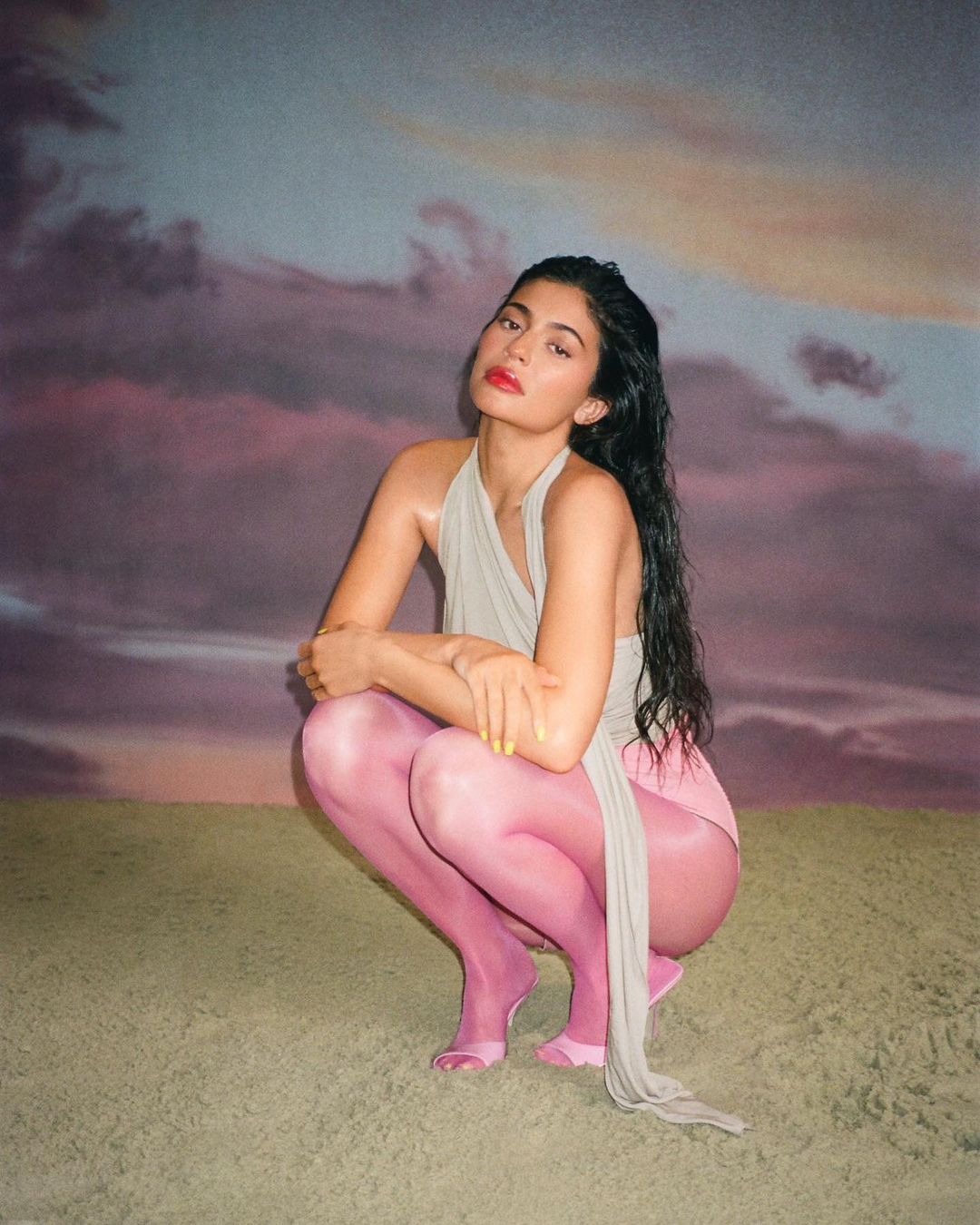 Woman with long, dark hair crouches in a studio setting, wearing a beige top, pink tights and matching toe shoes, posing against a dreamy background.