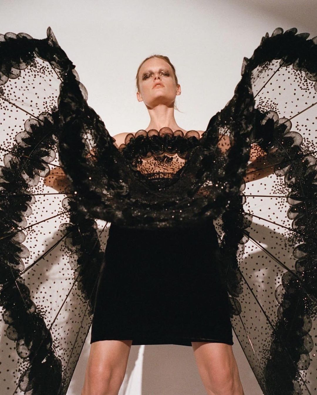 Fashion model poses in a black dress with elaborate black wings against a white background in a studio shot with high fashion makeup.