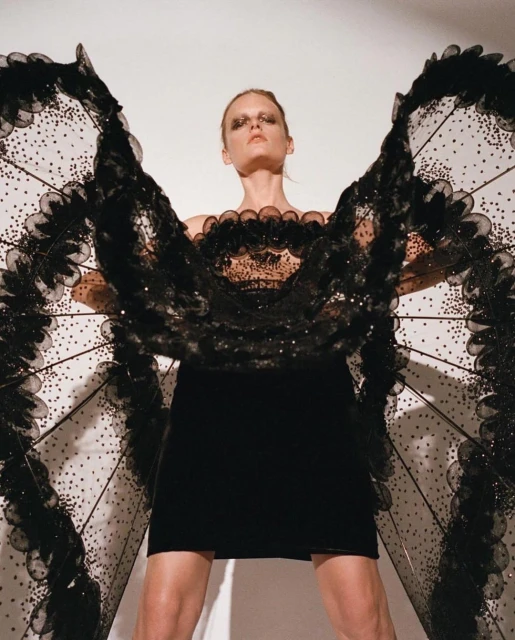 Fashion model poses in a black dress with elaborate black wings against a white background in a studio shot with high fashion makeup.