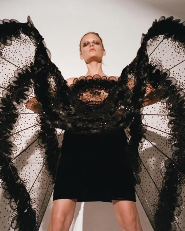 Fashion model poses in a black dress with elaborate black wings against a white background in a studio shot with high fashion makeup.