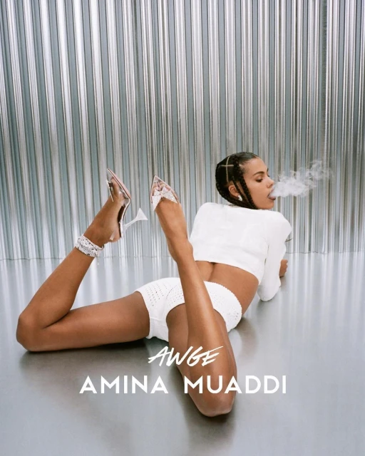 A woman exhales smoke while lying down in a white outfit and clear high heels, set against a corrugated metal backdrop.