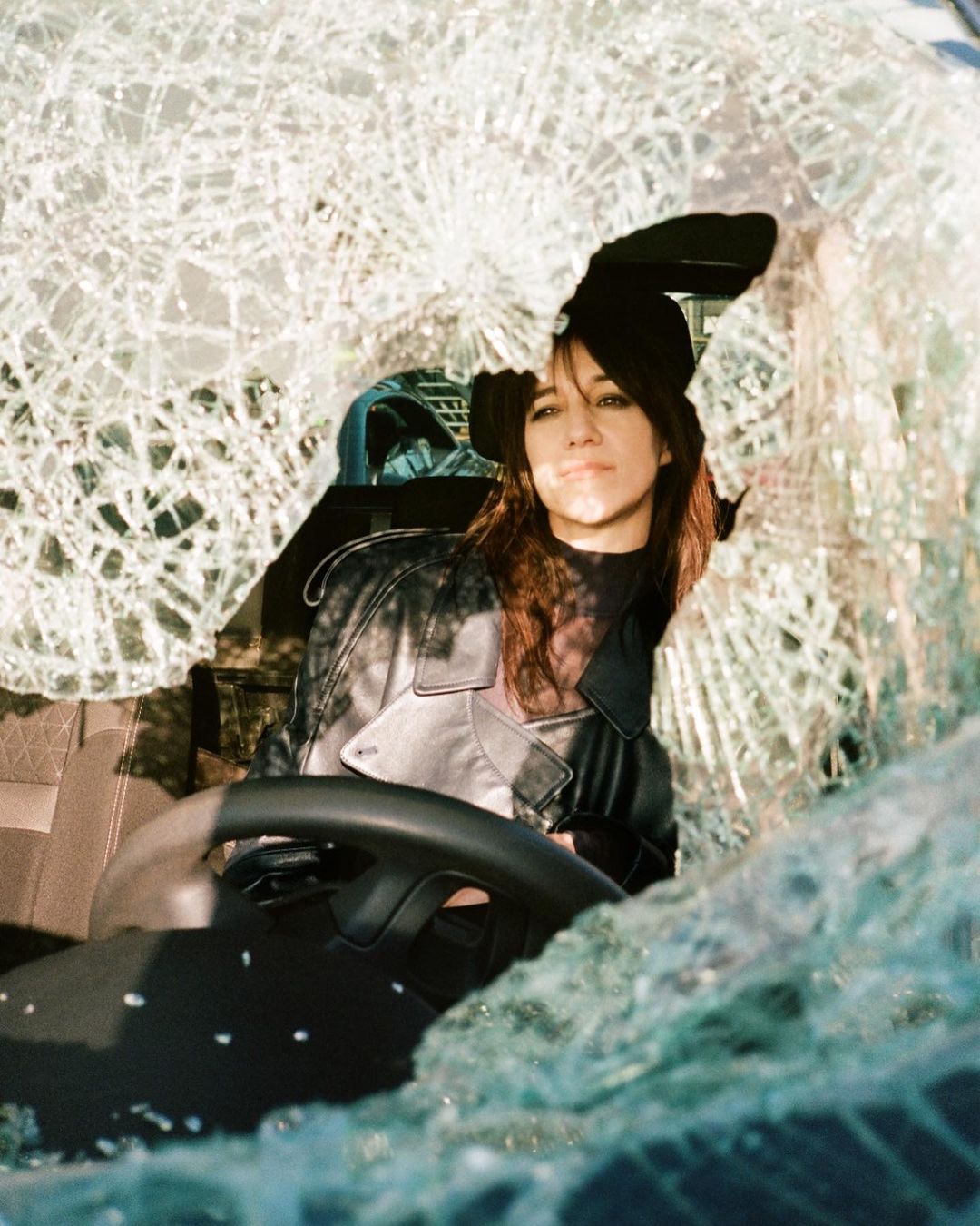 An adult female sits in a vehicle with a shattered windshield, wearing a leather jacket and hat. The scene is lit by daylight.