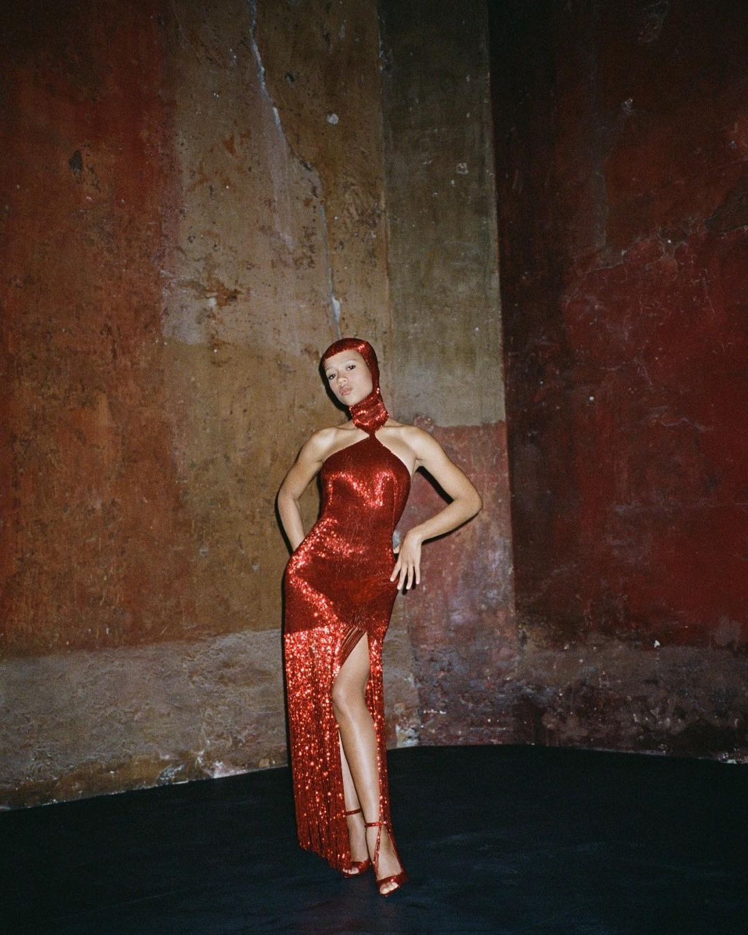 A woman in a red halter gown poses in an indoor space with textured walls, wearing matching strappy heels and head covering.