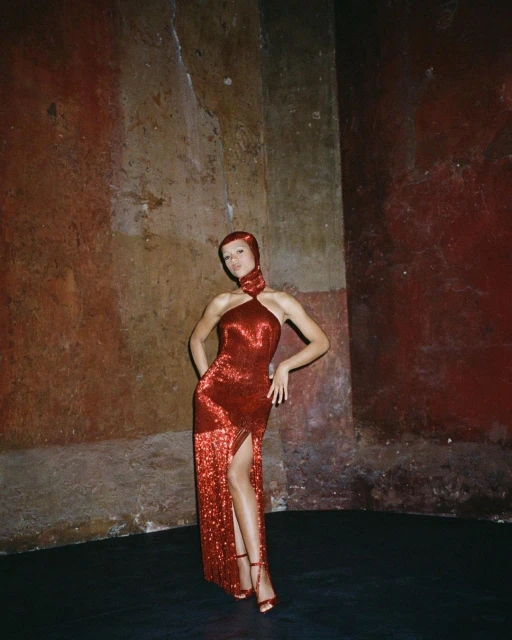 A woman in a red halter gown poses in an indoor space with textured walls, wearing matching strappy heels and head covering.