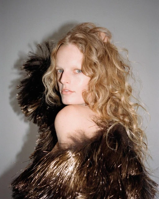 Blonde woman in gold fringe top poses against grey backdrop. Fashion portrait of a model wearing textured garment with soft lighting.
