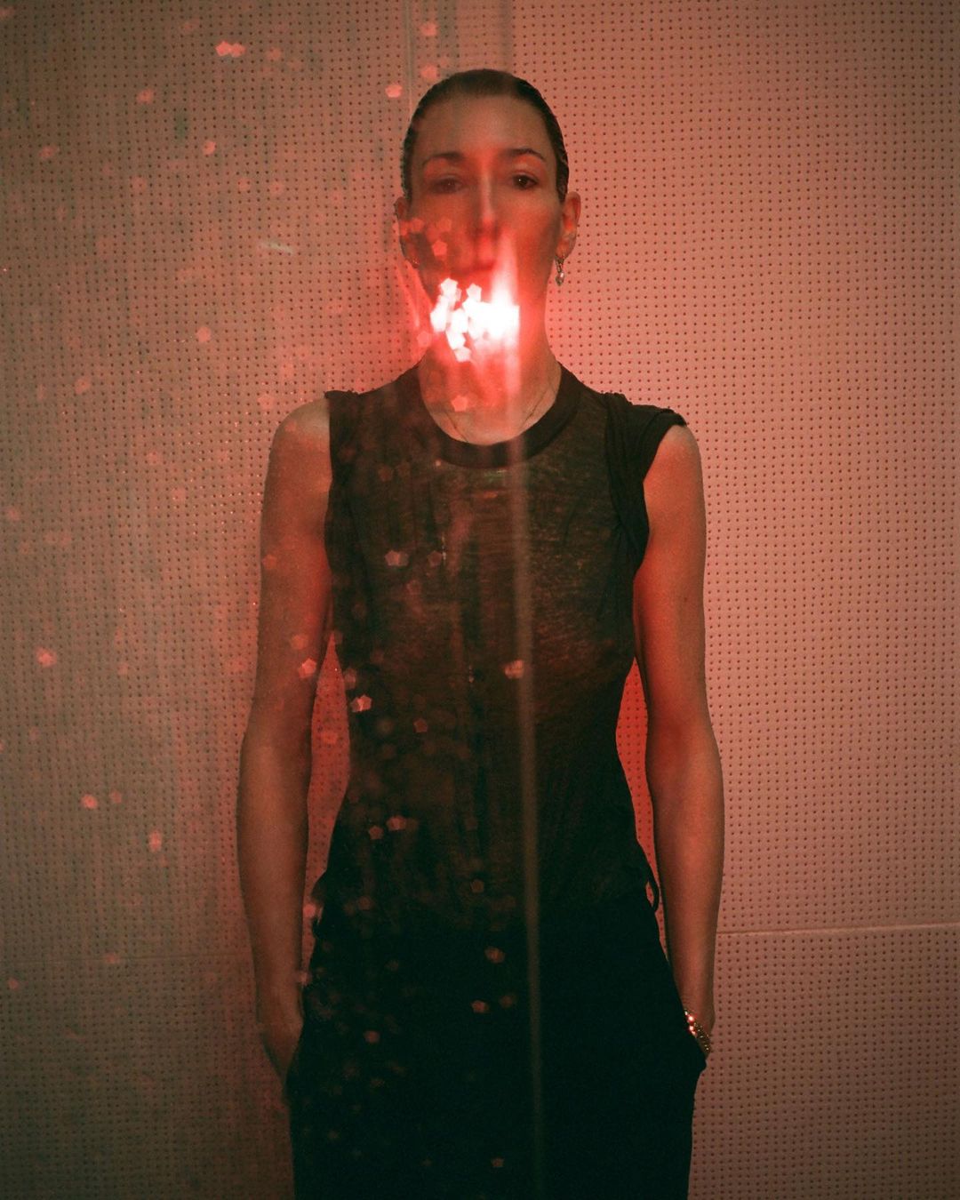 Portrait of a woman with red light and shadows on face and body, wearing a black top and pants in front of a textured wall.