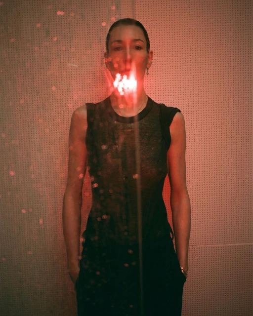 Portrait of a woman with red light and shadows on face and body, wearing a black top and pants in front of a textured wall.