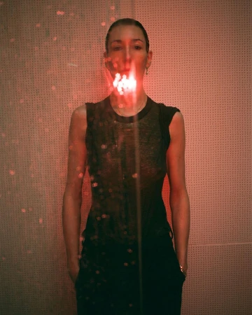Portrait of a woman with red light and shadows on face and body, wearing a black top and pants in front of a textured wall.