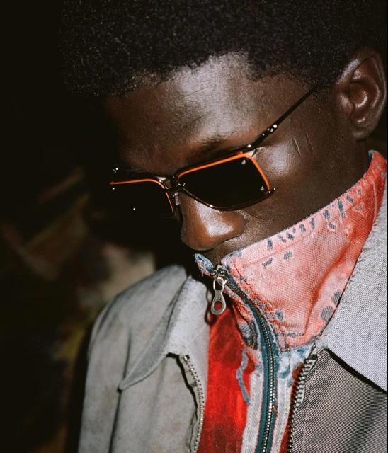 Close-up of a dark-skinned adult male with sunglasses, wearing a jacket and patterned shirt, in soft lighting against a dark backdrop.