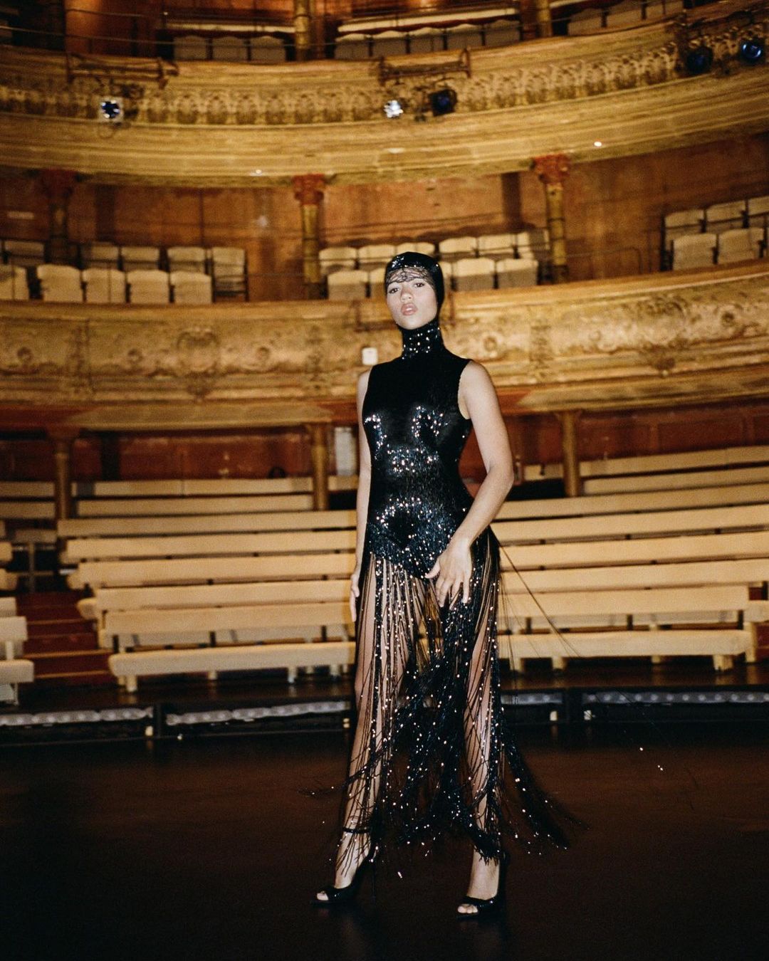 Elegant woman in a black sequined gown with fringe poses on a theater stage with rows of empty seating and ornate architectural details.
