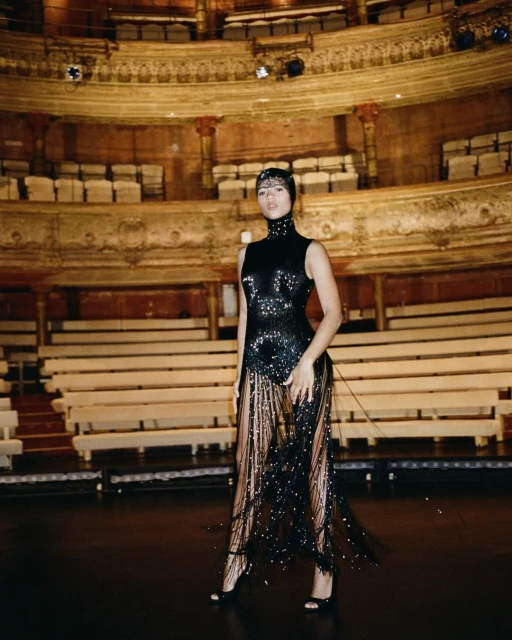 Elegant woman in a black sequined gown with fringe poses on a theater stage with rows of empty seating and ornate architectural details.