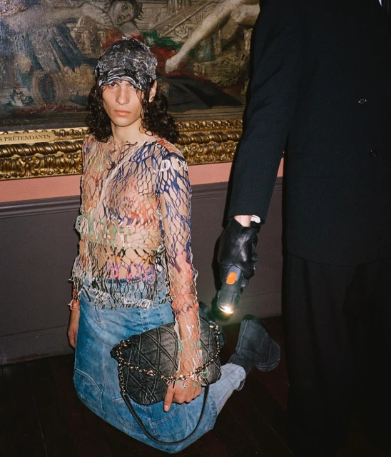 Person kneeling indoors next to another person holding a flashlight, wearing a netted top, jeans, hat, and handbag.