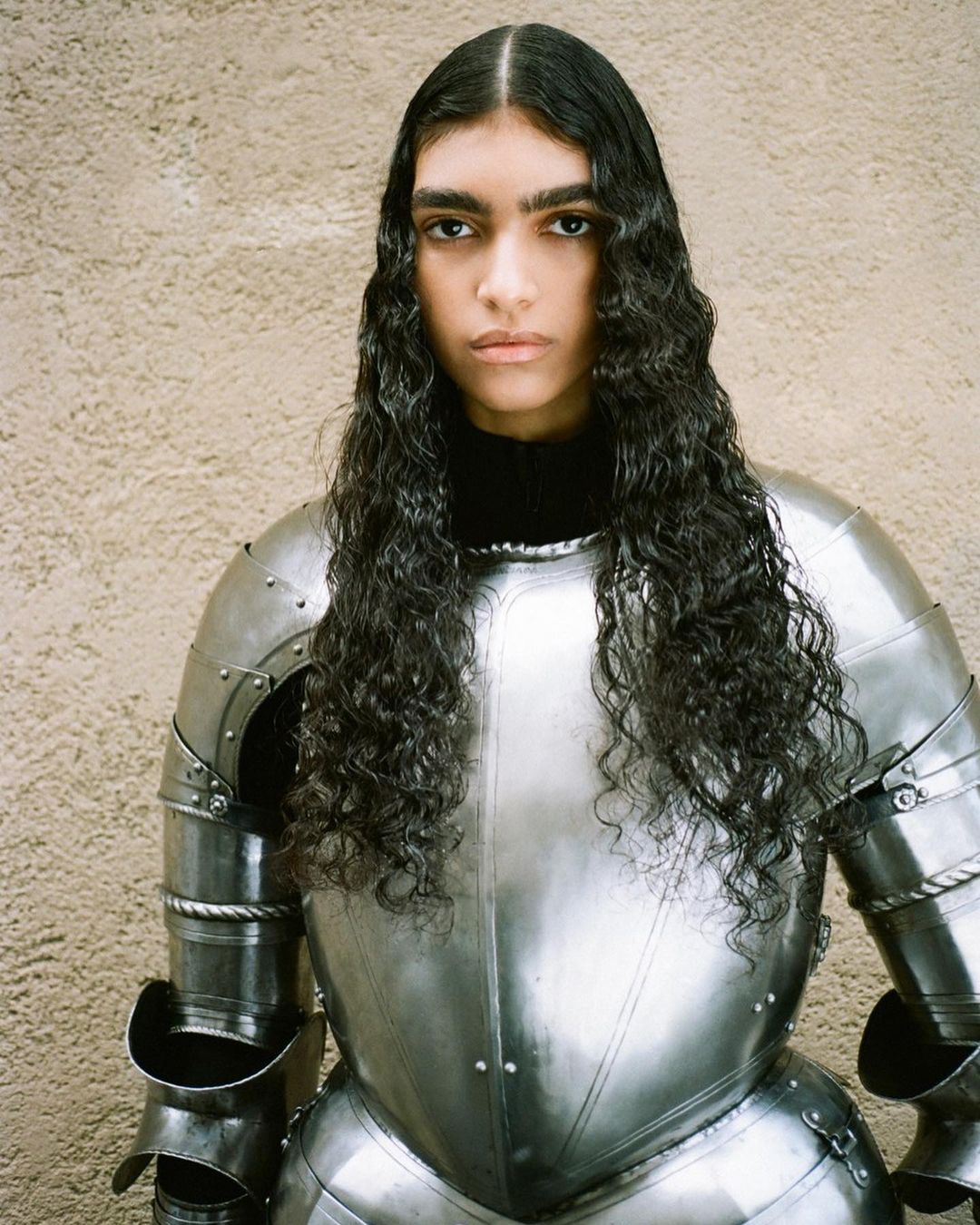Studio portrait of a woman with long, dark curly hair wearing reflective metal armor against a neutral beige background.