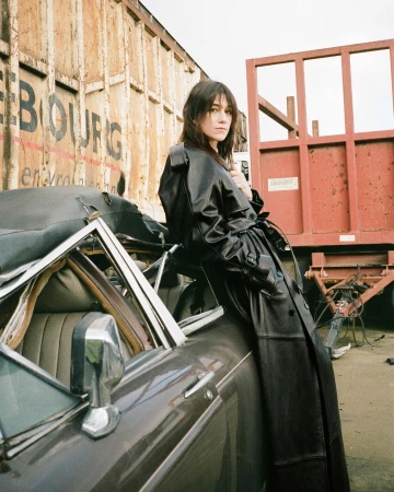 A woman in a black trench coat poses near an old car in a junkyard setting with a rail car container in the background.