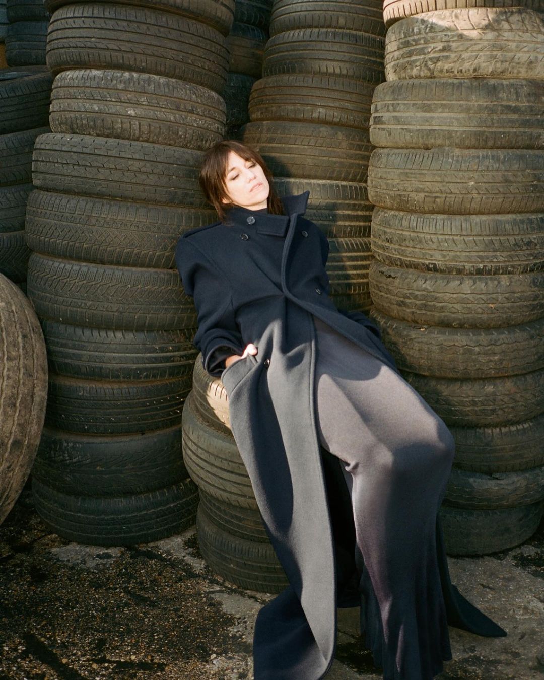 Adult female in an elegant dark coat and wide-legged trousers poses, leaning against a tall stack of car tires in outdoor setting.