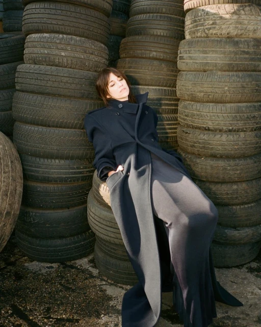 Adult female in an elegant dark coat and wide-legged trousers poses, leaning against a tall stack of car tires in outdoor setting.