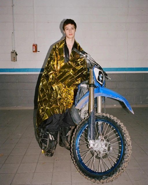 A young adult stands with a motocross bike, draped in a gold foil blanket, set against a simple brick wall in an indoor garage.