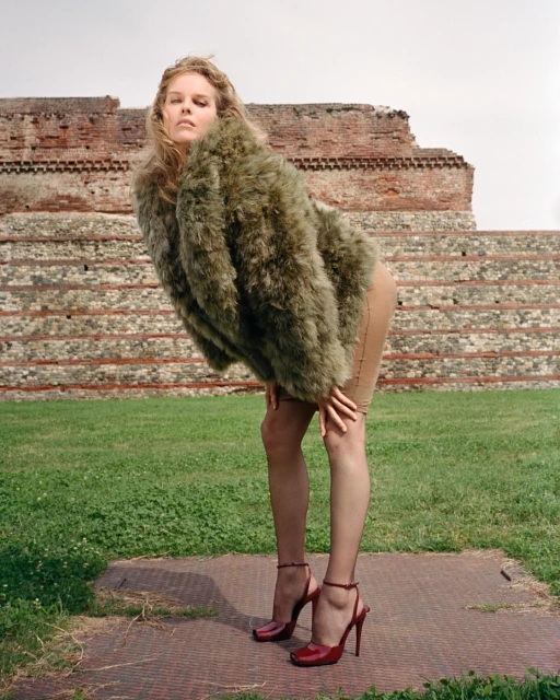 A young woman in a fur coat and red heels poses in a grassy field in front of an old brick wall in an outdoor fashion shot.