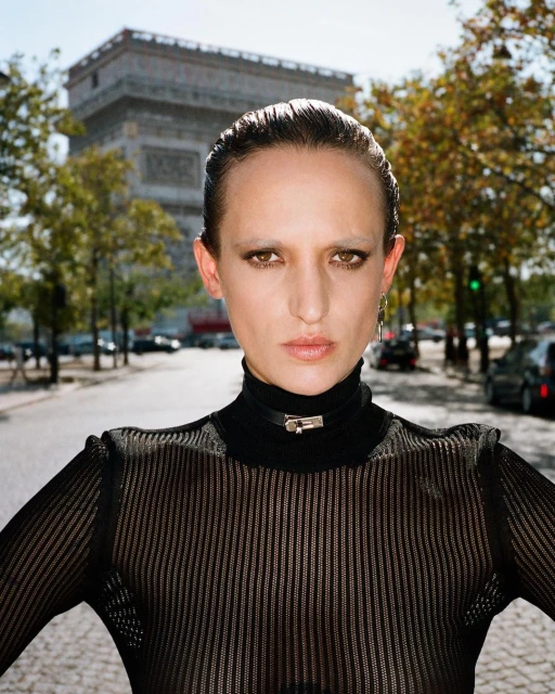 Woman with slicked-back hair and sheer top stands in Paris with Arc de Triomphe visible in the background on a sunny day.