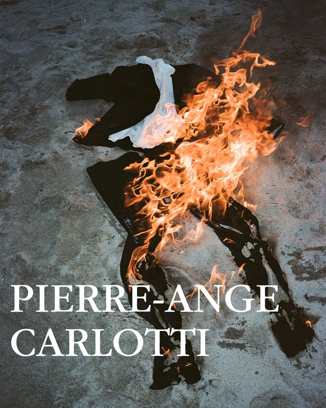 Burning black suit with a white shirt on a sandy surface, engulfed in bright orange flames, conveying a dramatic and destructive scene.