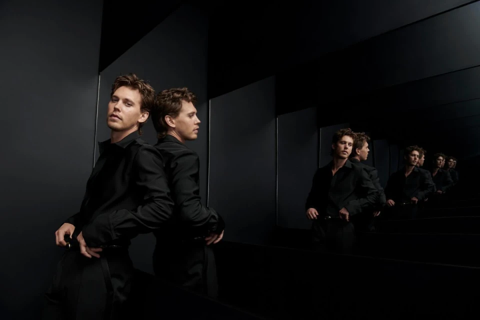 Man in a black shirt poses, reflected in a dark mirror, creating a receding series of images in an indoor, high-contrast setting.