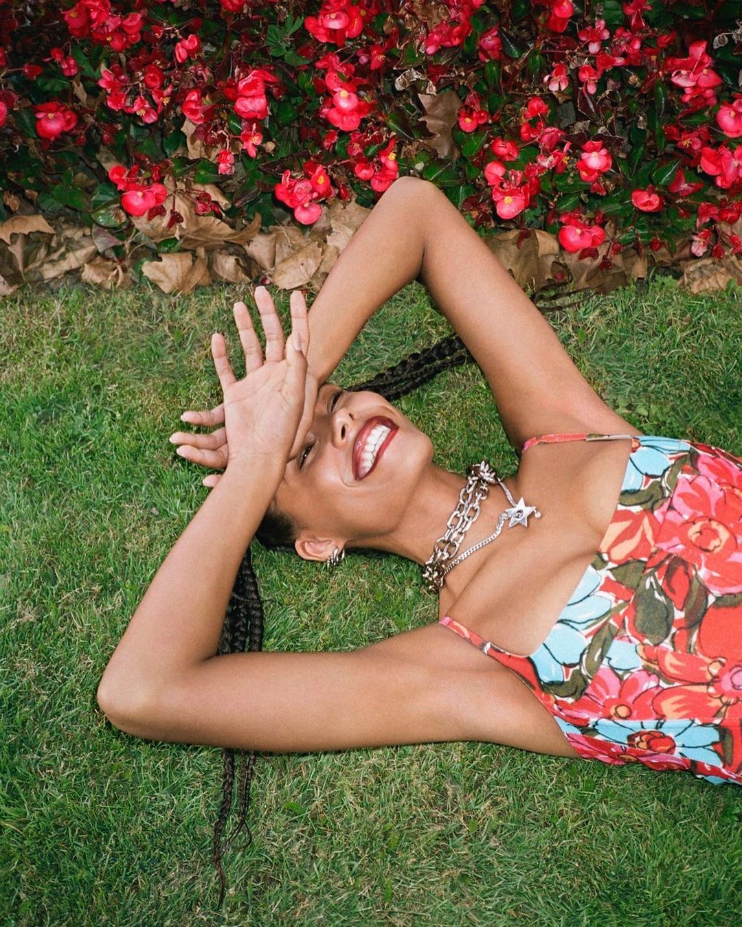 Smiling woman with braided hair lying in green grass wearing a floral dress and silver chain necklace